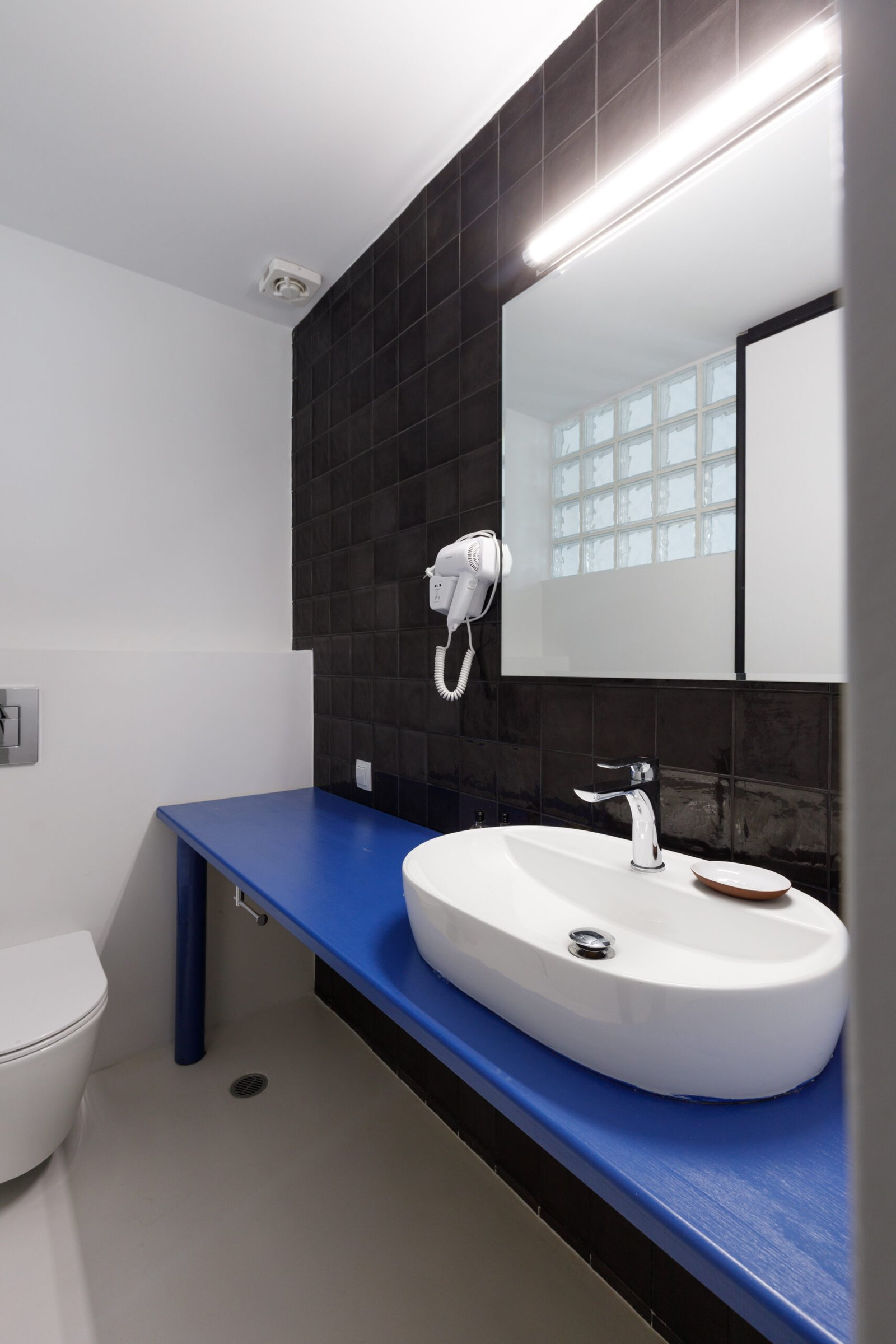bathroom of baroness 2, with black wall tiles, a big mirror, white sink on blue countertop and a toilet